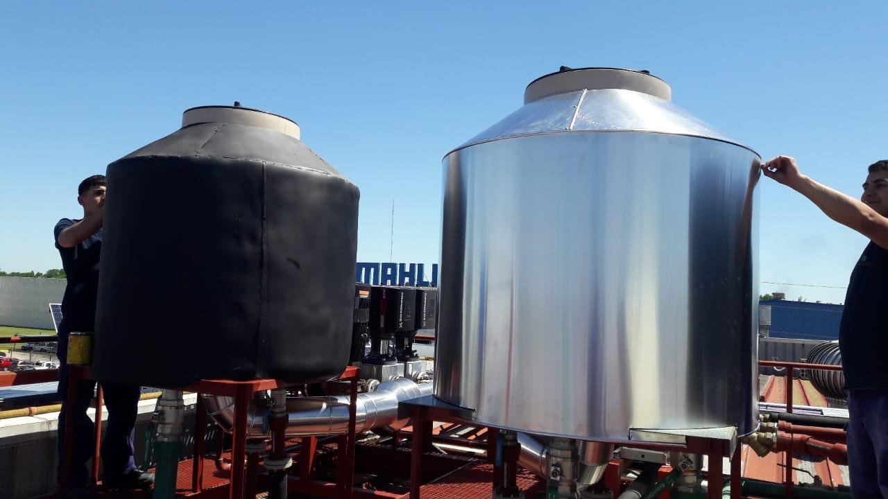 Dos tanques de agua, uno cubierto con una funda negra y otro de acero inoxidable, en la azotea de un edificio, con dos hombres trabajando en él, bajo un cielo despejado.