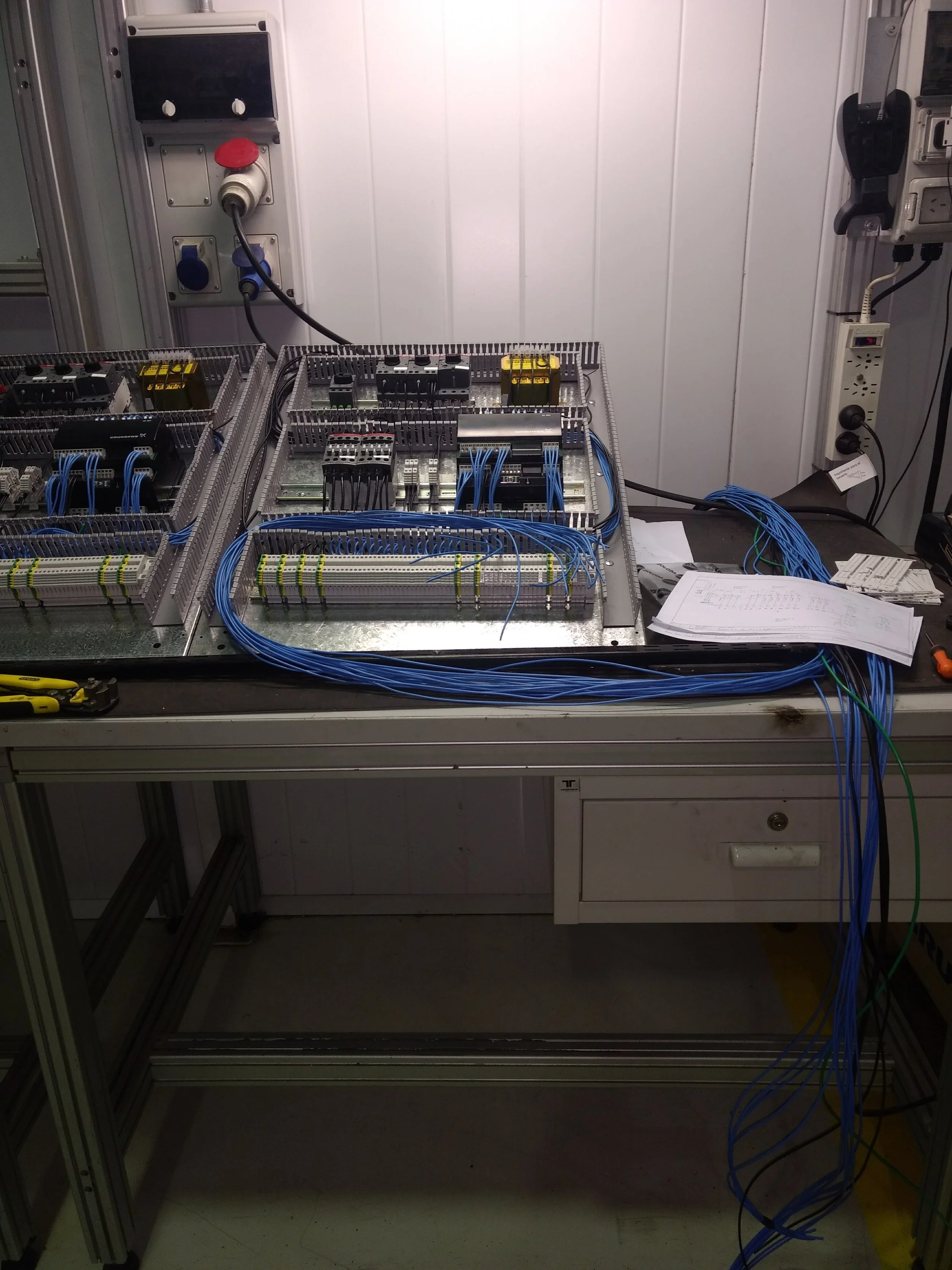 Cuadro de control eléctrico con cables azules, cajas, herramientas y papeles en un taller técnico.