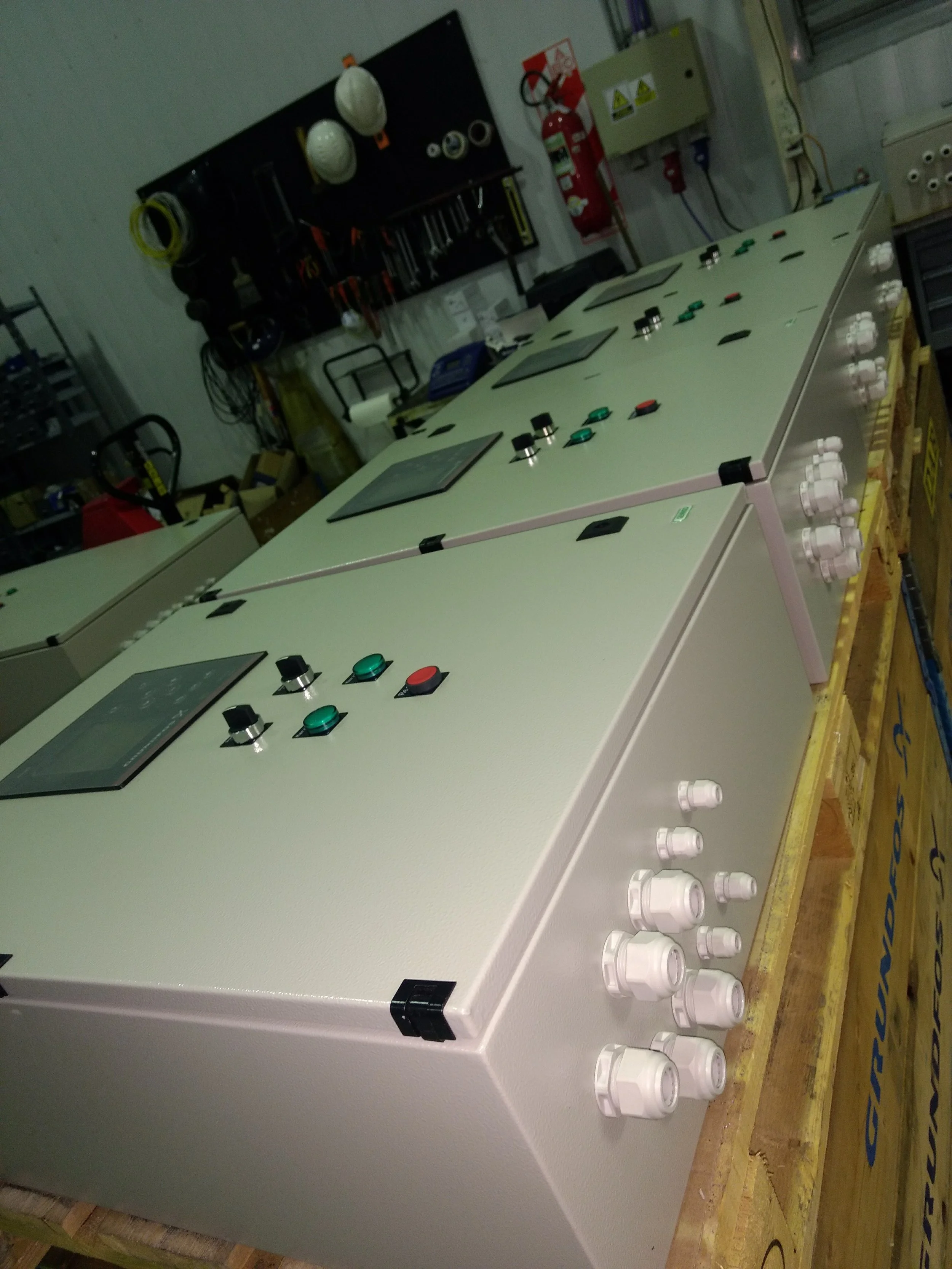 Una mesa de control con botones y pantallas en un ambiente de taller o laboratorio, con cables, cascos de seguridad, extintor y otros equipos en las paredes y estanterías.