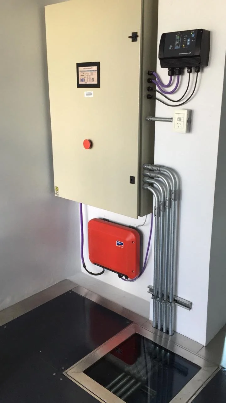 Caja eléctrica de color beige con pantalla digital y botón rojo, instalada en la pared junto a un controlador con cables de color morado y negro, cables de metal que bajan por la pared, una caja roja en la parte inferior y un tapete negro en el suelo