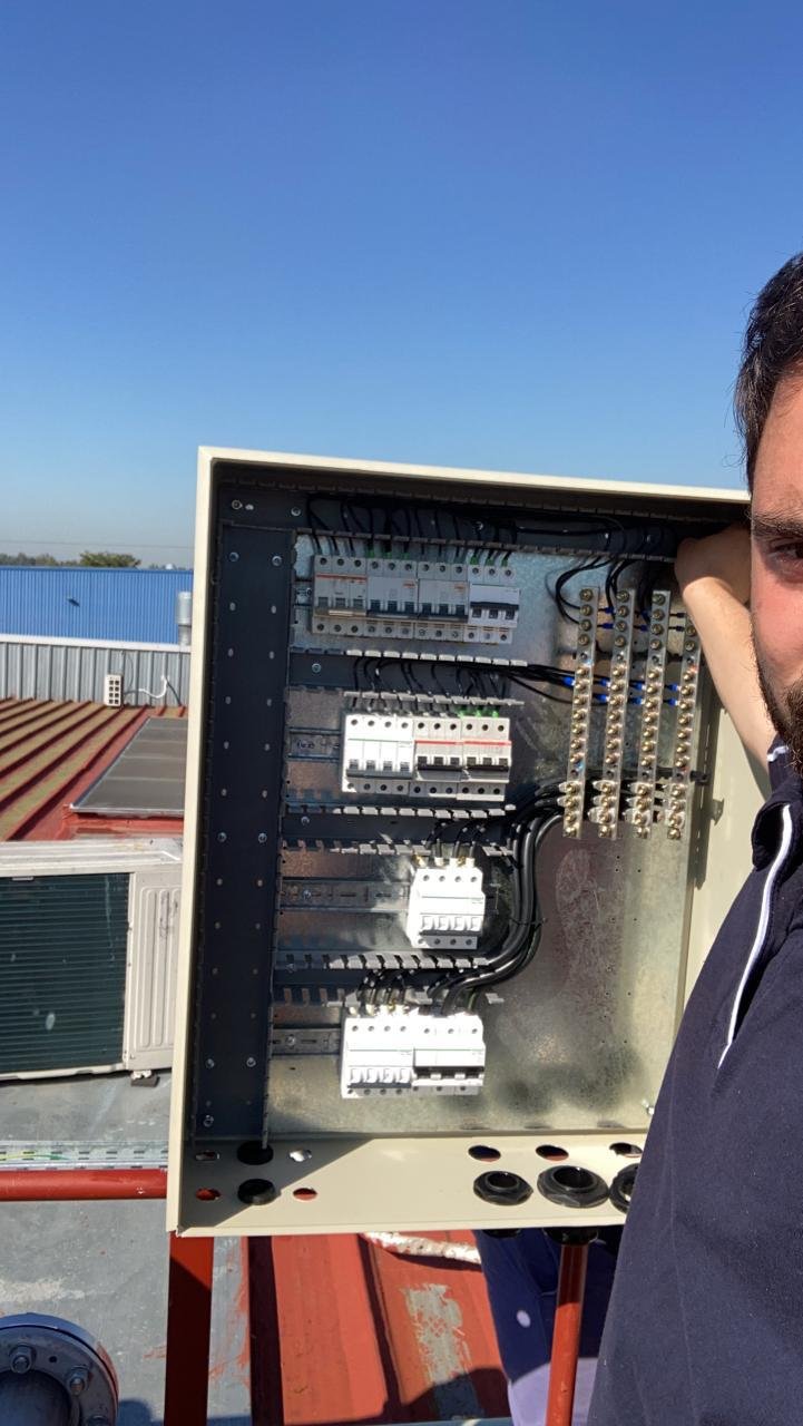 Hombre sujetando un tablero de control eléctrico con componentes y cables, en un techo con cielo despejado.