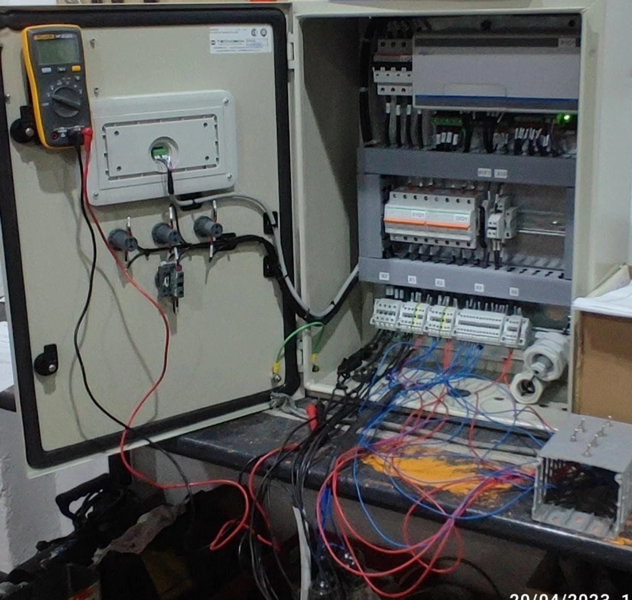 Caja de control eléctrico con componentes y cables conectados, equipo de medición y multímetro a la izquierda.
