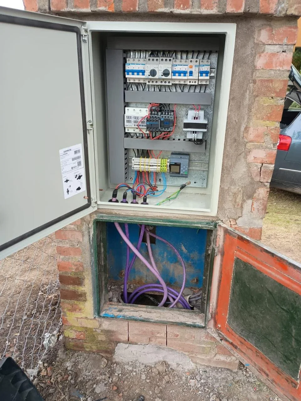 Caja de distribución eléctrica con cables y componentes eléctricos en una pared de ladrillos, con puerta abierta y cables morados debajo.