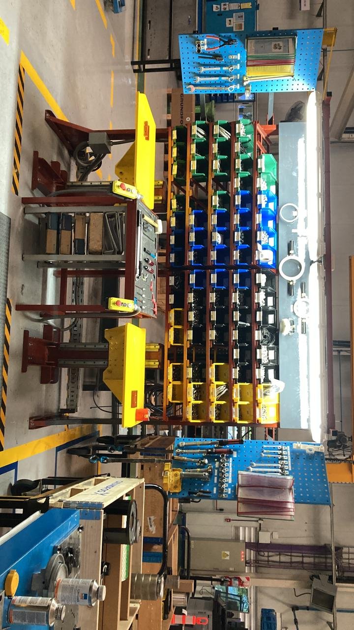 Área de trabajo en una fábrica con estantes de colores y herramientas, incluyendo un máquina en primer plano y un tablero de herramienta azul en el fondo.