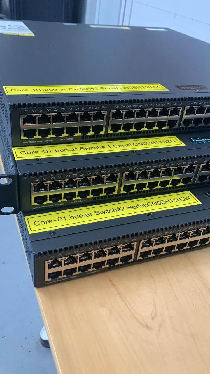 Tres switches de red apilados sobre una mesa, con etiquetas amarillas que indican sus nombres y números de serie.