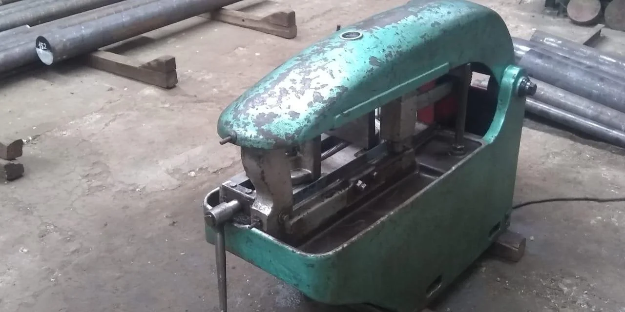 Máquina de corte o calibrado industrial de metal, de color verde, en un taller de fabricación, con piezas de metal y madera alrededor.