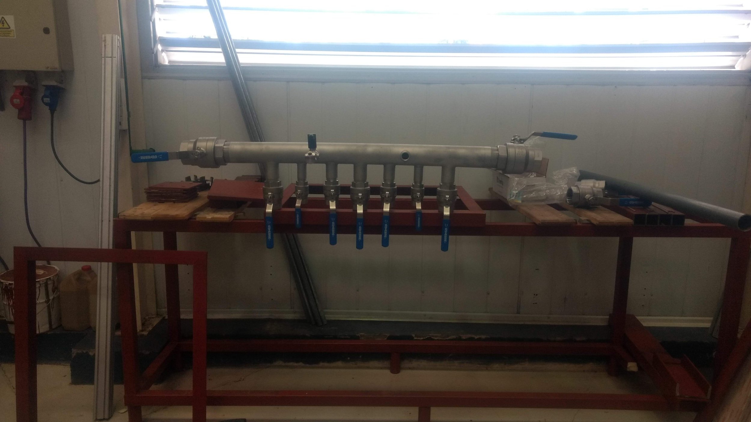 Conjunto de tuberías de metal con válvulas en una mesa de trabajo, cerca de una ventana con persianas horizontales.