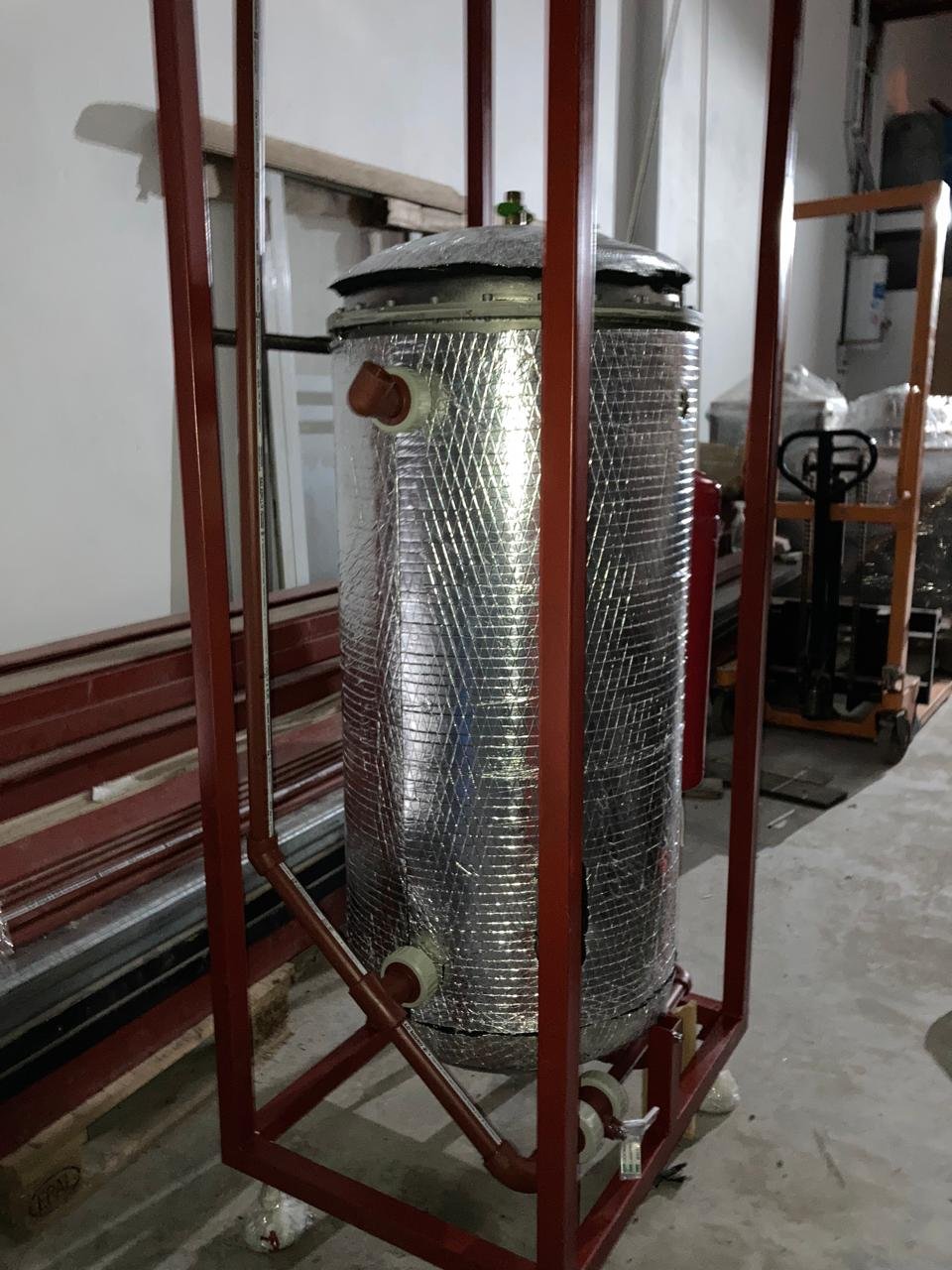 Un tanque de agua aislado con un revestimiento de fibra de vidrio, instalado dentro de una estructura metálica roja en un espacio industrial o de almacenamiento.
