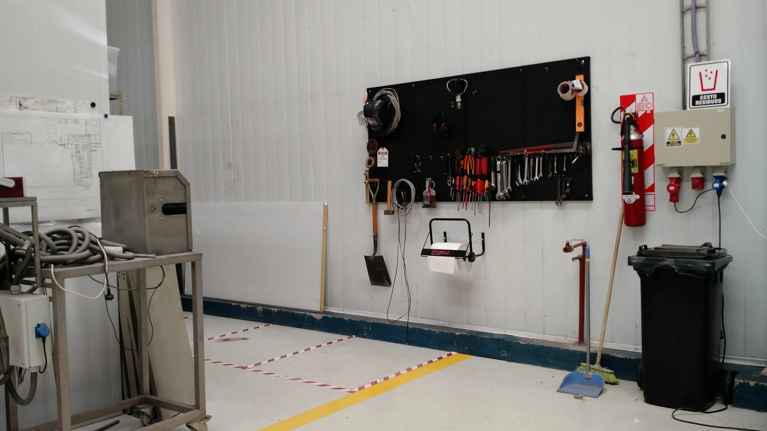 Herramientas y equipo de limpieza en un taller, con un panel de herramientas en la pared, extinguidores, y una zona delimitada en el suelo.