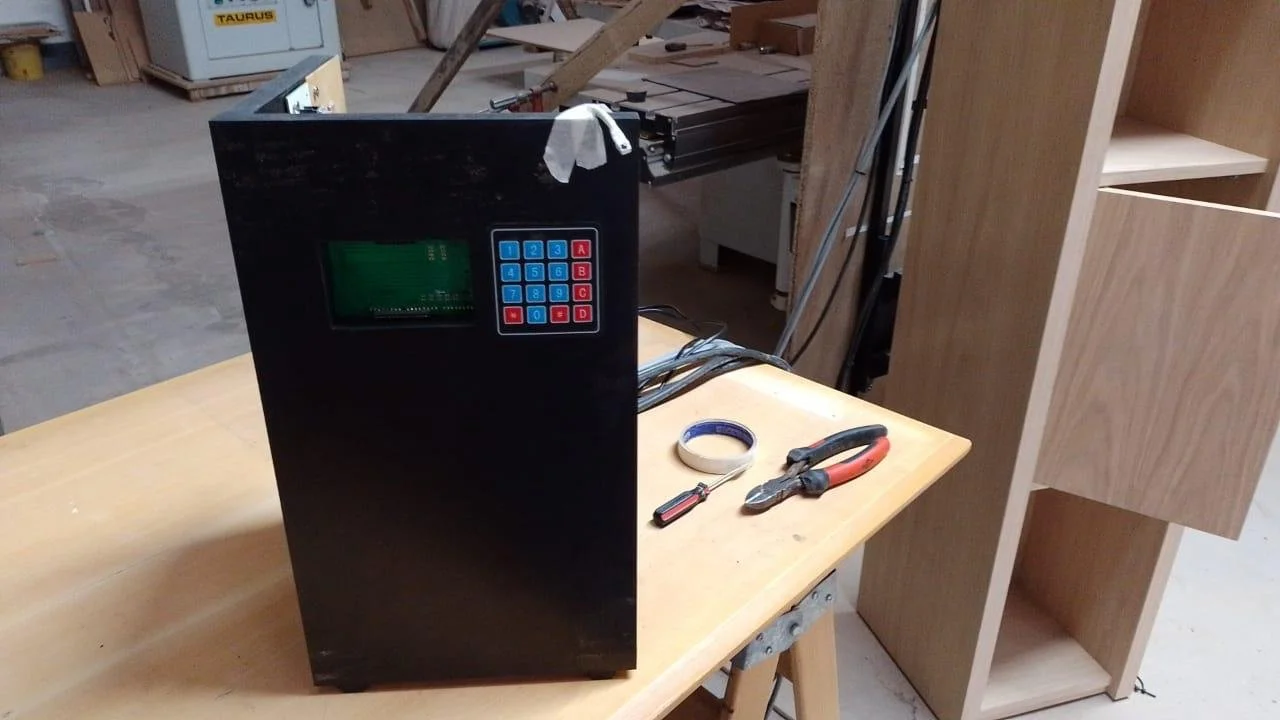 Dispositivo electrónico negro de caja rectangular con pantalla y teclado numérico en un taller de carpintería rodeado de herramientas y madera