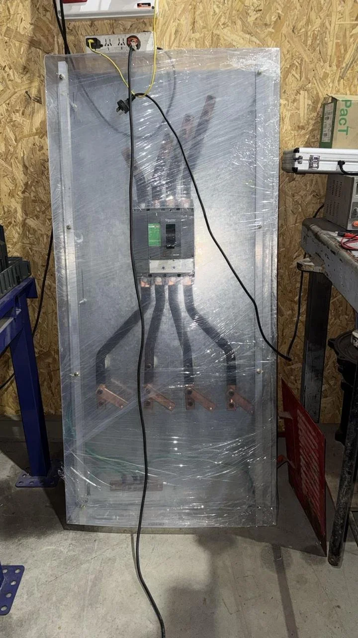 Un panel eléctrico con varias conexiones eléctricas, cubierto por una lámina de plástico transparente, en un taller de construcción con paredes de madera