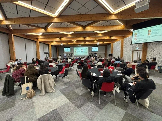 A large conference room filled with attendees seated at round tables, facing a stage with multiple screens displaying a presentation.