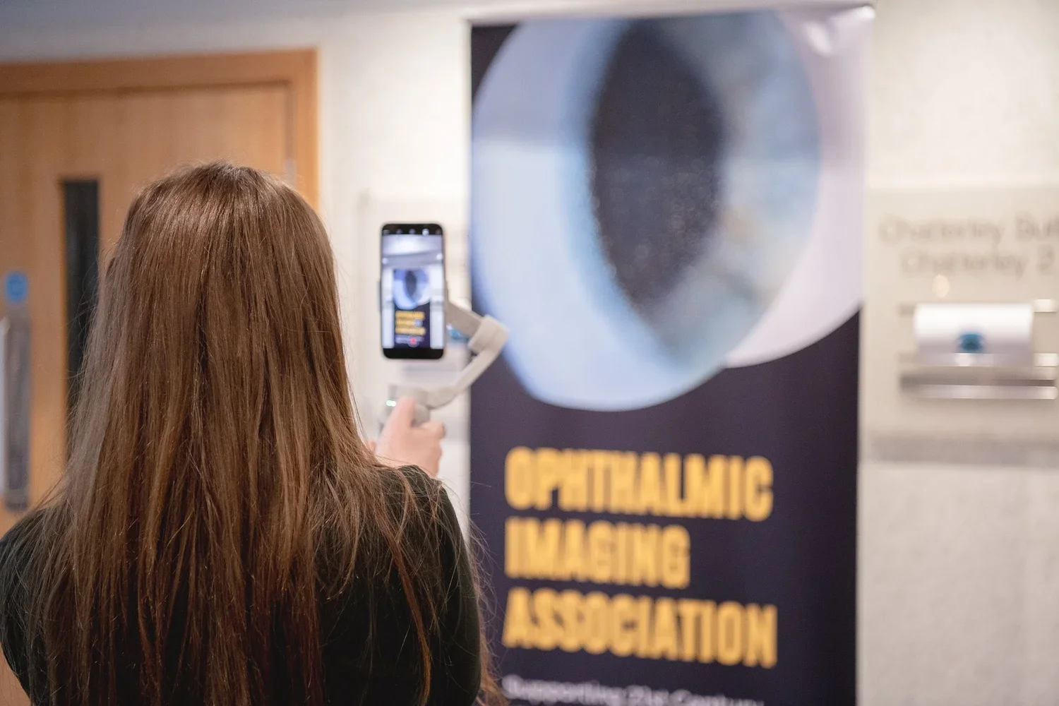 A person with long brown hair taking a selfie with a smartphone on a selfie stick in front of a poster that has an eye graphic and the text 'OPHTHALMIC IMAGING ASSOCIATION'.