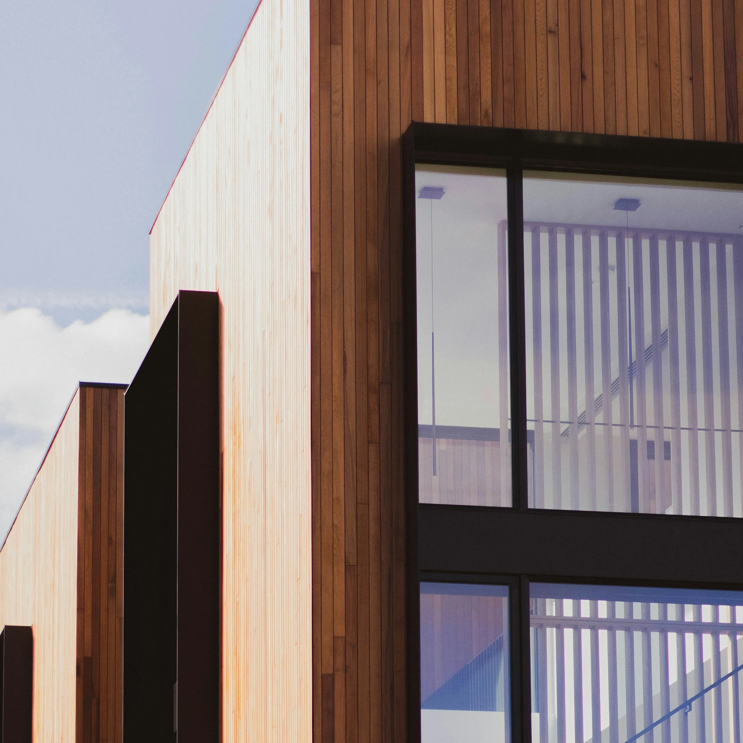 Close-up view of a modern building facade with wooden paneling and large glass windows, reflecting a blue sky and neighboring structures.