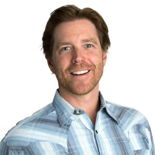A smiling man with reddish-brown hair, a beard, and blue eyes, wearing a light blue striped button-down shirt, against a white background.