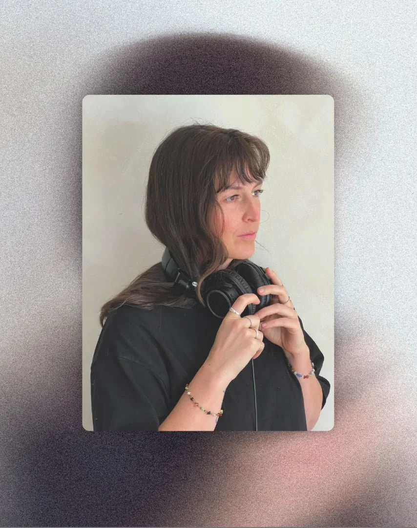 Une jeune femme avec des cheveux bruns portant un t-shirt noir, tenant un casque audio noir autour du coup, avec une expression pensive, devant un fond blanc.