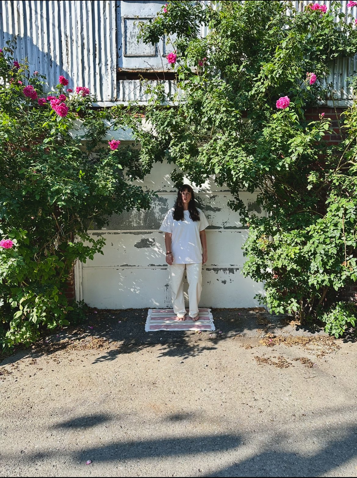 Une femme debout sur un tapis à rayures, portant des vêtements blancs, sous un arbre fleuri avec des roses roses, devant un mur blanc usé et une porte en métal.