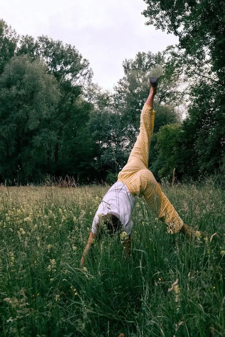 Une personne faisant une acrobatie en équilibre sur une main dans un champ herbeux avec des arbres en arrière-plan.