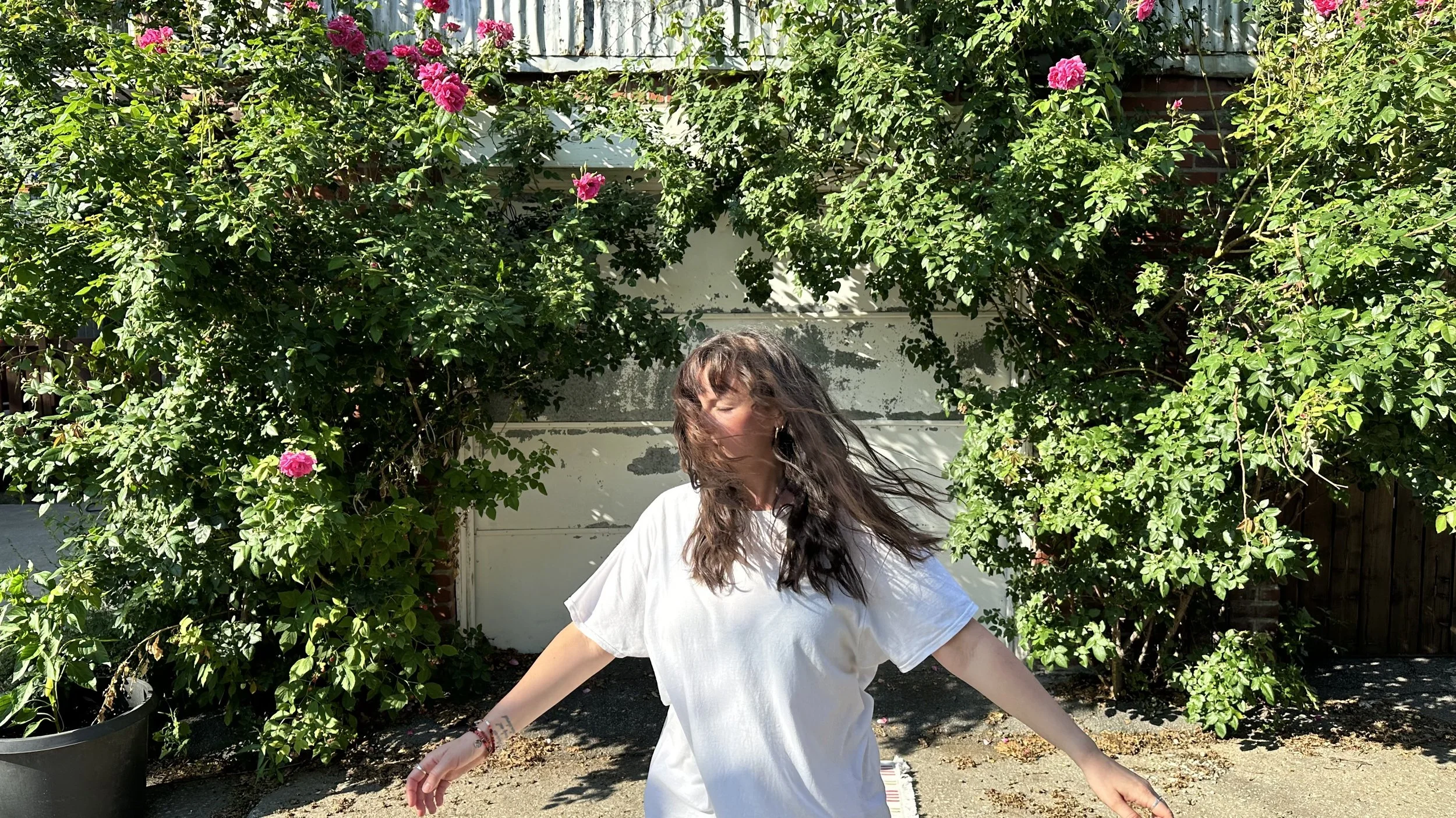 Fille aux cheveux longs et ondulés portant une robe blanche, dansant ou sautant devant un mur recouvert de feuillage vert avec des fleurs roses
