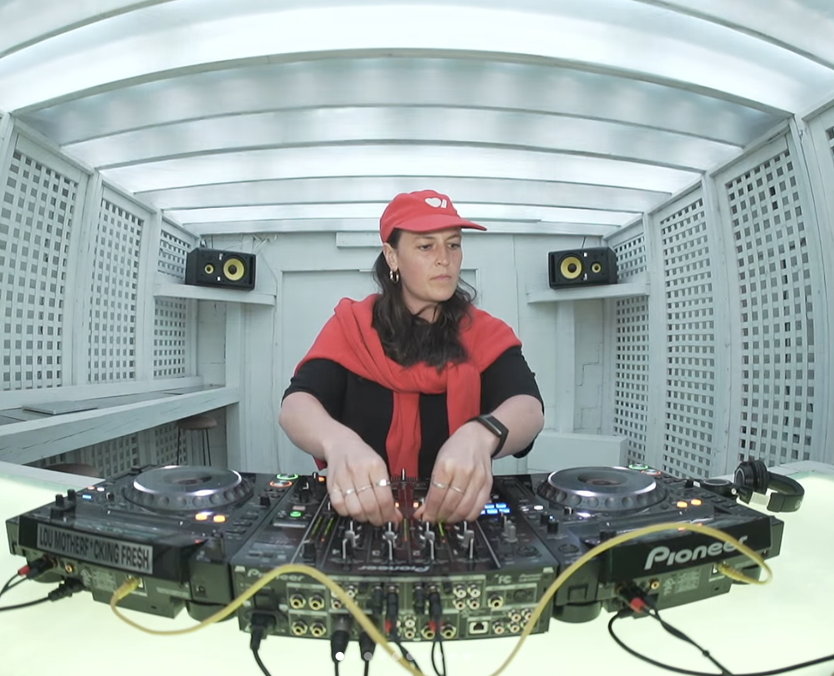 DJ femme en train de mixer dans une cabine, portant une casquette rouge et un foulard rouge, avec deux enceintes noires en arrière-plan.
