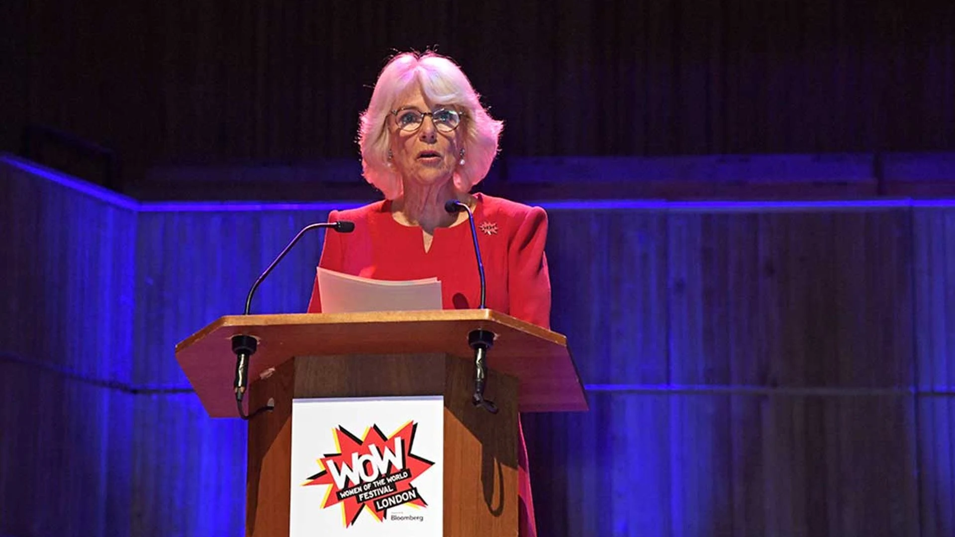 An elderly woman with white hair, glasses, and a red blazer speaking behind a podium at the Women of the World Festival in London. The podium has a logo that says "WOW Women of the World Festival London".