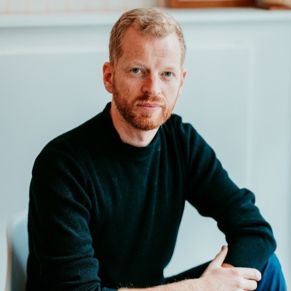 A man with red hair and a beard, wearing a black long-sleeve shirt, sitting at a table with a neutral expression in a light-colored room.