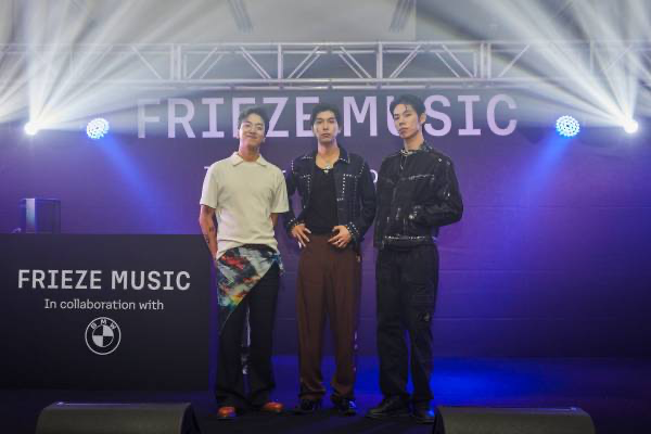 Three men standing on a stage in front of a large screen with the words "FREIZE MUSIC" during an event or concert, with lighting effects and a DJ booth to the left that displays the Frieze Music logo and a BMW brand logo.