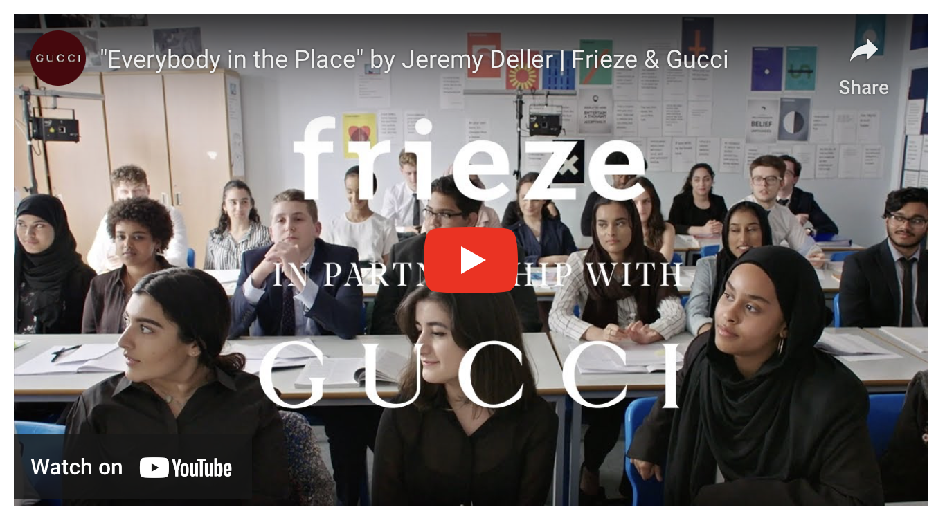 A classroom filled with diverse students sitting at desks, listening attentively, with a large overlay text promoting 'Everybody in the Place' by Jeremy Deller, Frieze, Gucci, and a YouTube link.