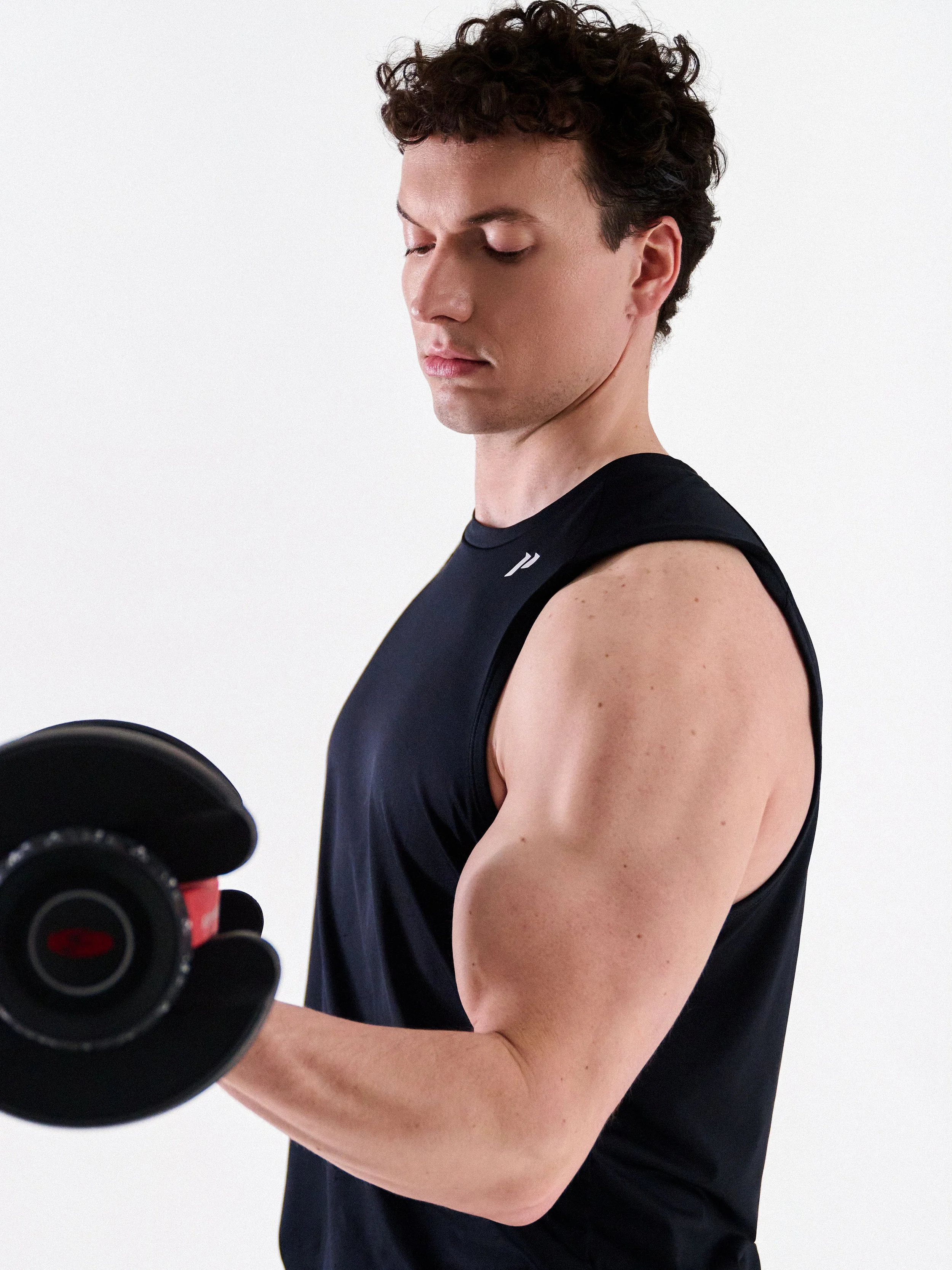 A young man with short curly hair lifting a dumbbell, wearing a black sleeveless athletic shirt, against a plain white background.