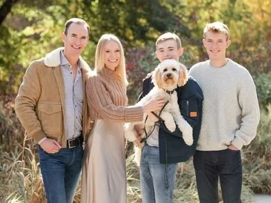 Two parents and two teen boys stand in the sunshine, dressed in shades of beige and blue, and one boy holds a white fluffy dog