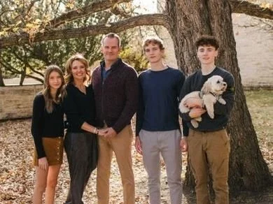 Family of five stands in a wooded area dressed in brown, navy, and black. A teenage boy holds a small white dog.