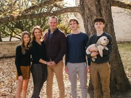Family of four standing outdoors by a large tree, with a man, woman, and two teenage boys, one of whom is holding a small dog.