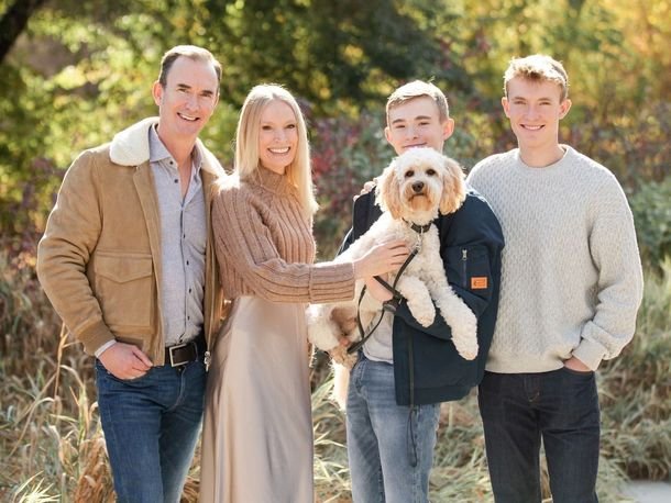 Family of four outdoors in autumn holding a small dog, all smiling at the camera.