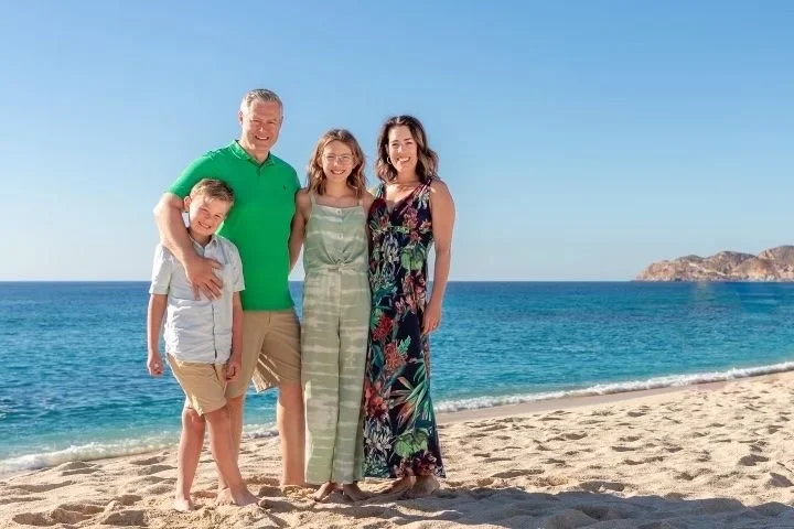 Family of four standing on the beach with the ocean and clear sky in the background, smiling and dressed in casual summer clothing.