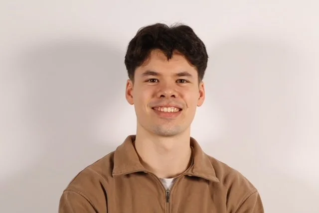 Young man with dark curly hair wearing a brown sweater, smiling 