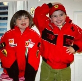 Two children dressed in Ferrari racing team jackets, smiling indoors.