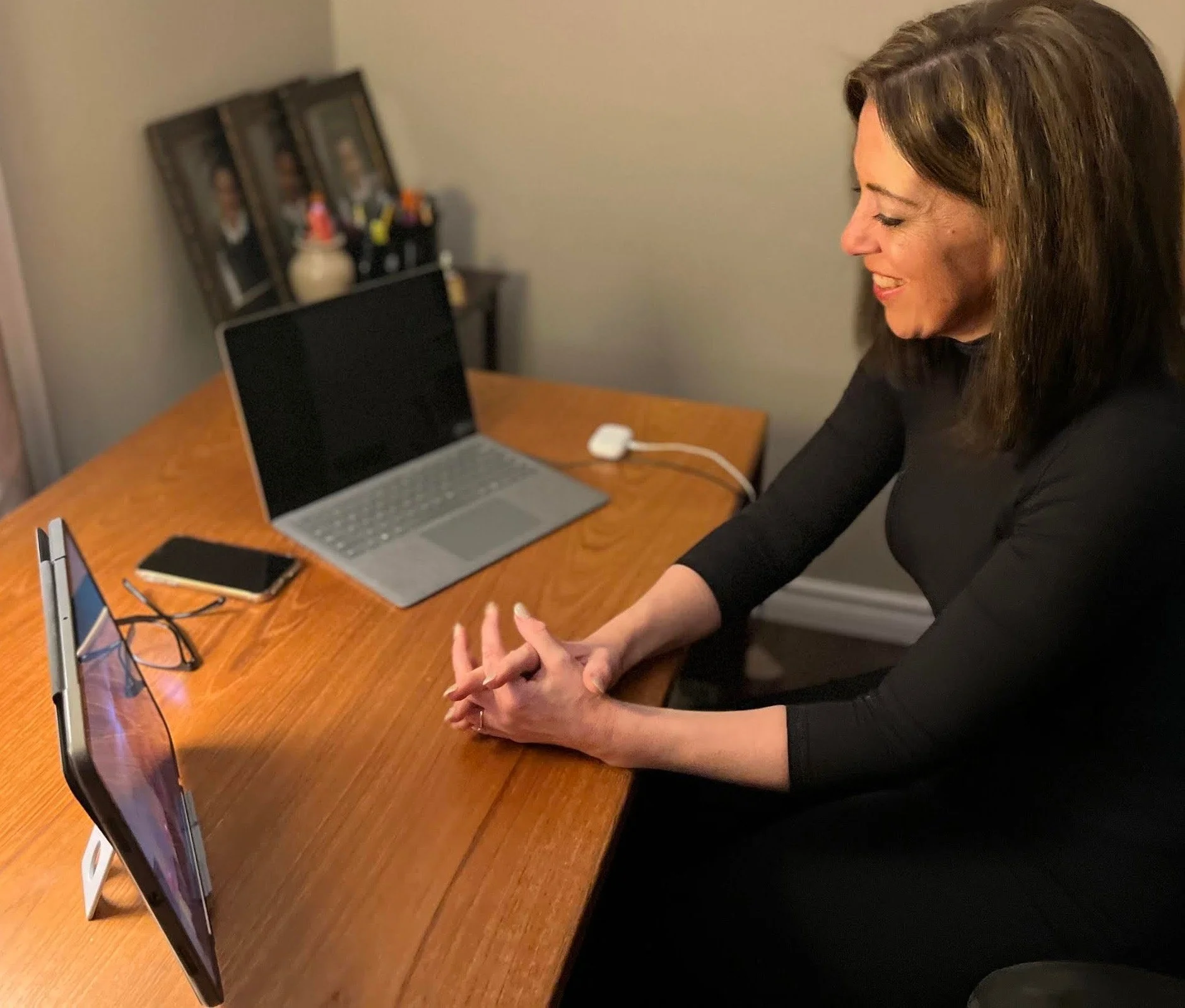 A woman with brown hair and a black top sitting at a wooden table, smiling, with a laptop, a smartphone, and a display stand with paper and a pen on the table, and framed photographs.