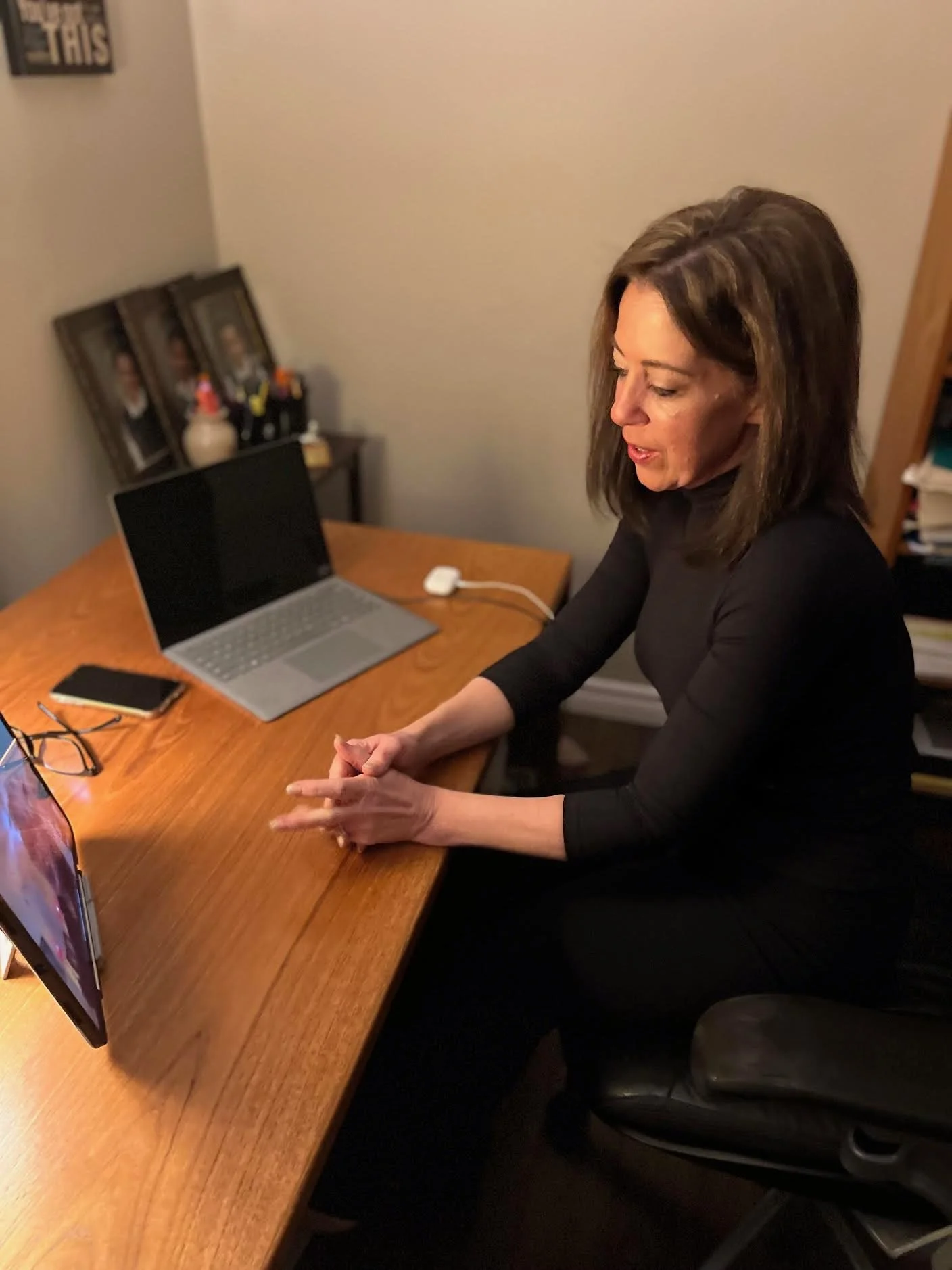 A woman sitting at a wooden desk with a laptop, notebook, and glasses, looking at her phone. Behind her are framed photographs and a small collection of figurines or decorations. The room has beige walls and a bookshelf with books.