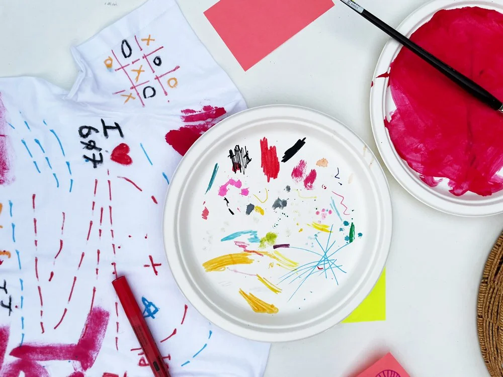 A white paper plate with colorful paint smears and lines. A small white towel with red, black, and blue marker drawings and writing, including a red heart. Several paintbrushes and paper scraps are also visible, suggesting an art or craft activity.