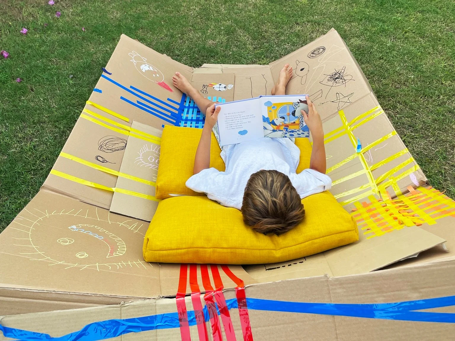 Child lying on a yellow pillow inside a large cardboard box decorated with colorful tape, drawing and writing on the box with markers, and reading a picture book outdoors on grass.