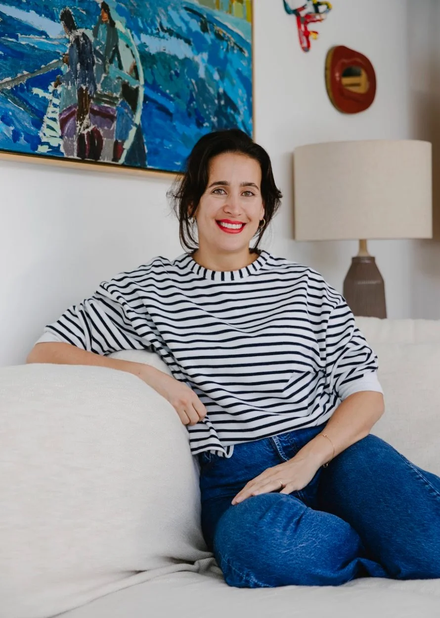 A woman with dark hair, red lipstick, and a striped shirt sitting on a white couch in a living room.