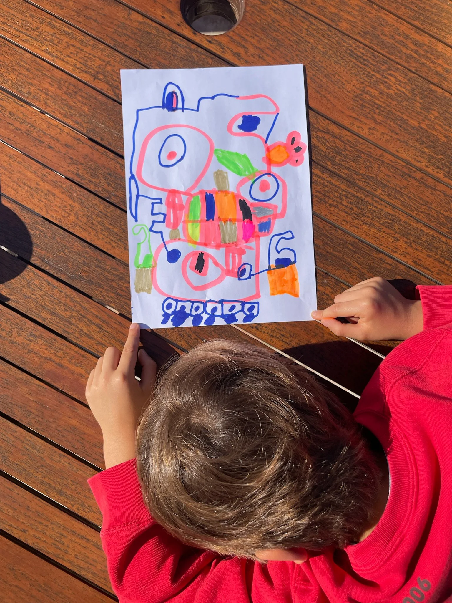 A child with brown hair wearing a red long-sleeve shirt, sitting at a wooden table, holding a colorful drawing with both hands, with the drawing rotated