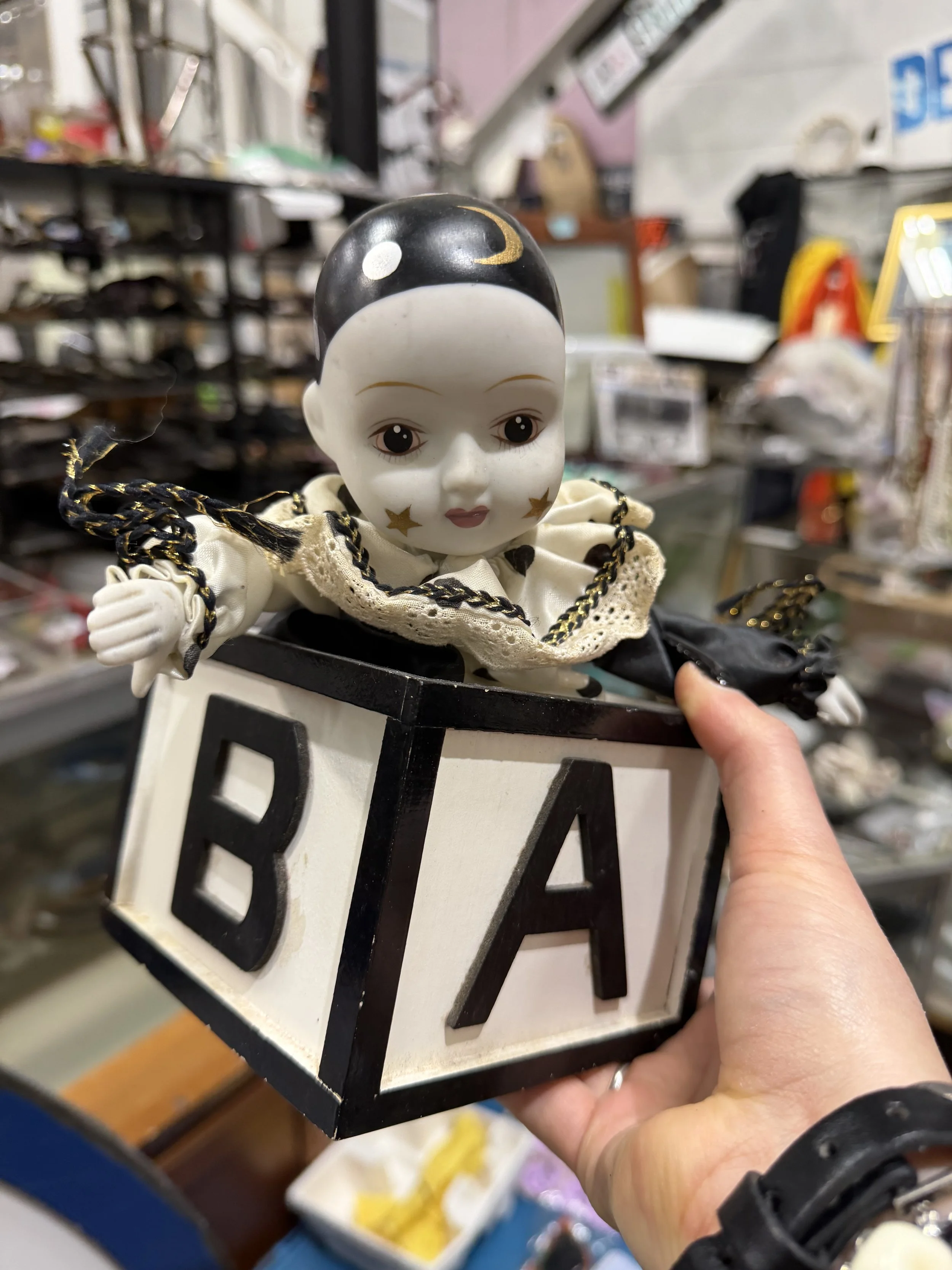 A person holding a white and black decorative doll with a smiling face, stars on cheeks, wearing lace and embroidery details, inside a small black and white box with cut-out pattern, in a cluttered store with various objects on shelves in the background.
