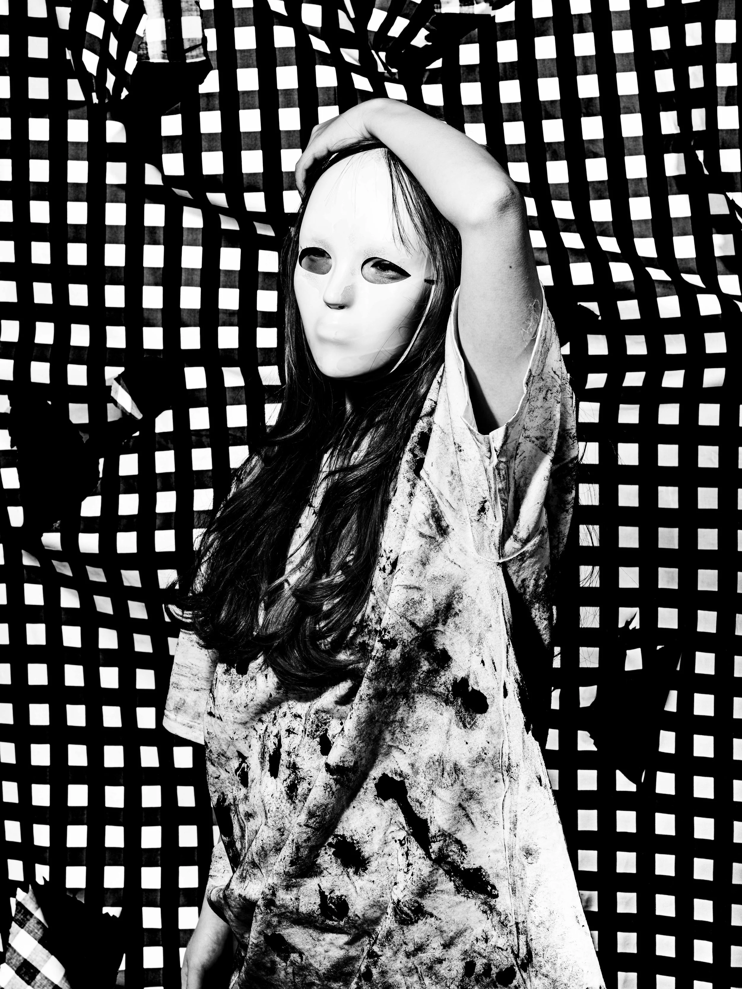 A person wearing a mask with no facial features, standing in front of a checkered backdrop. The person's long hair is visible, and they are dressed in a paint-splattered shirt or robe. Their right hand is raised, touching their head.