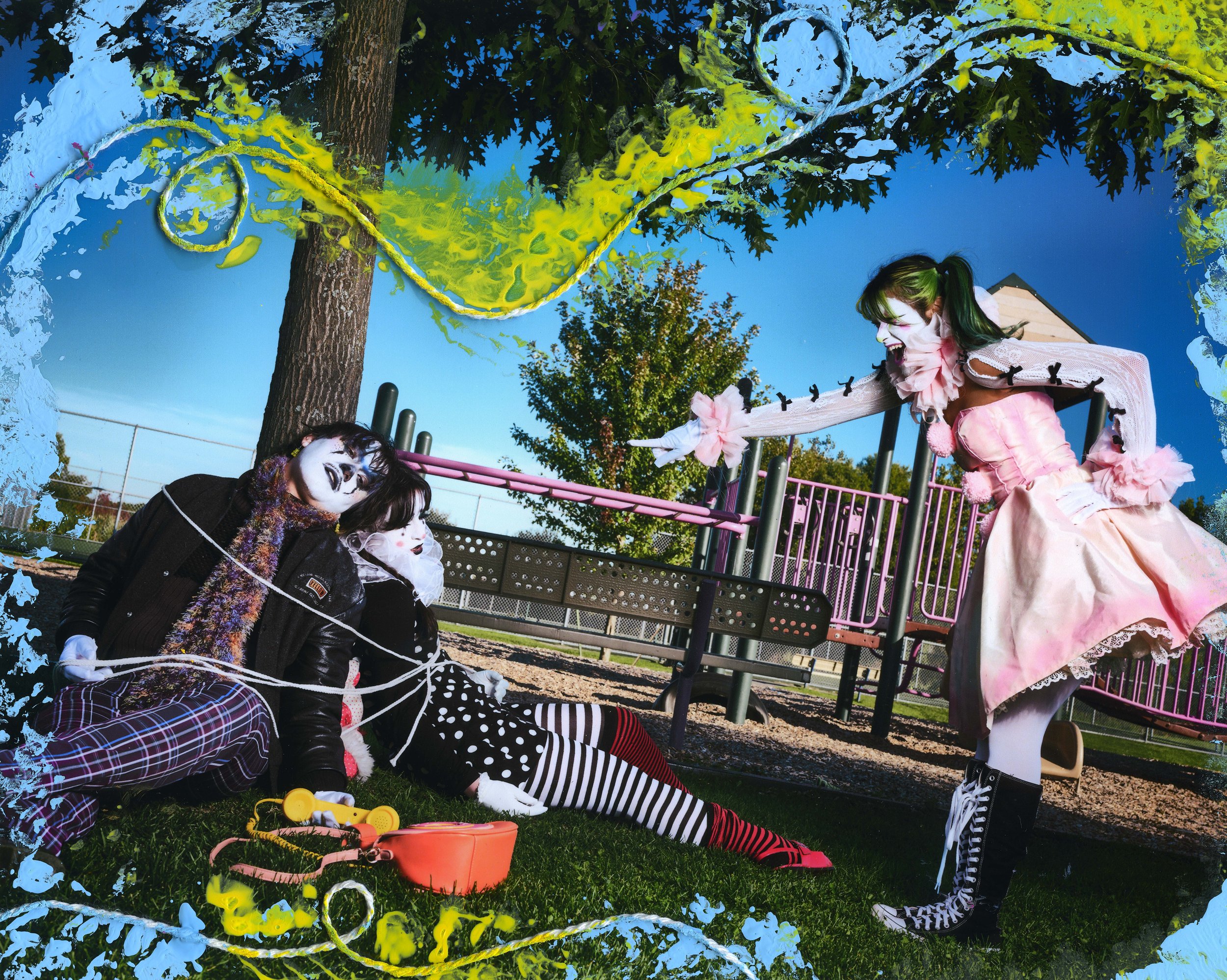 A group of people dressed in clown costumes and makeup at an outdoor playground, with some sitting on the ground and one standing near a pink railing, under a tree on a sunny day.