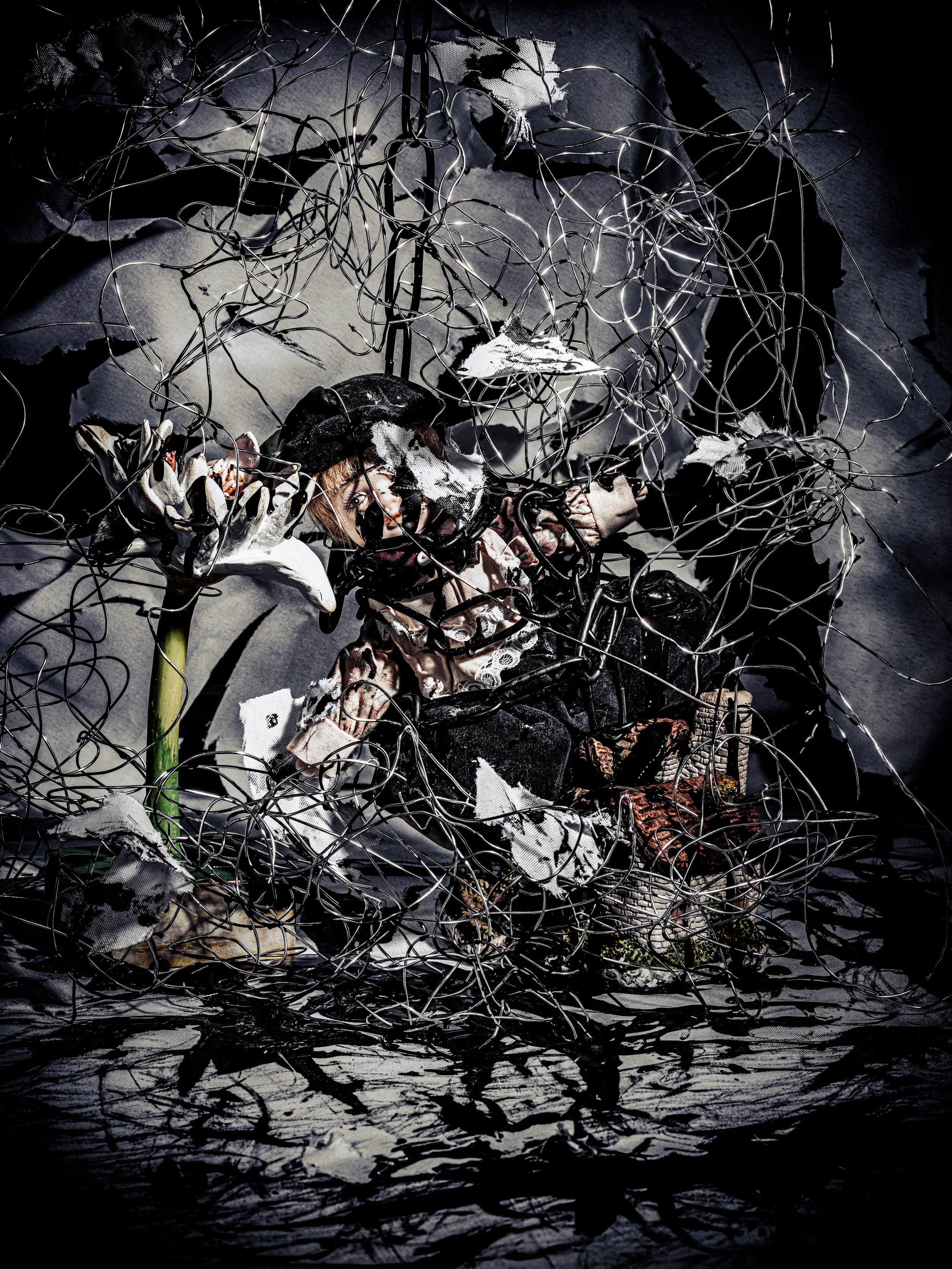 An art installation with tangled wires, torn fabric, a mannequin with a painted face, and flowers, set against a dark background with a reflective surface.