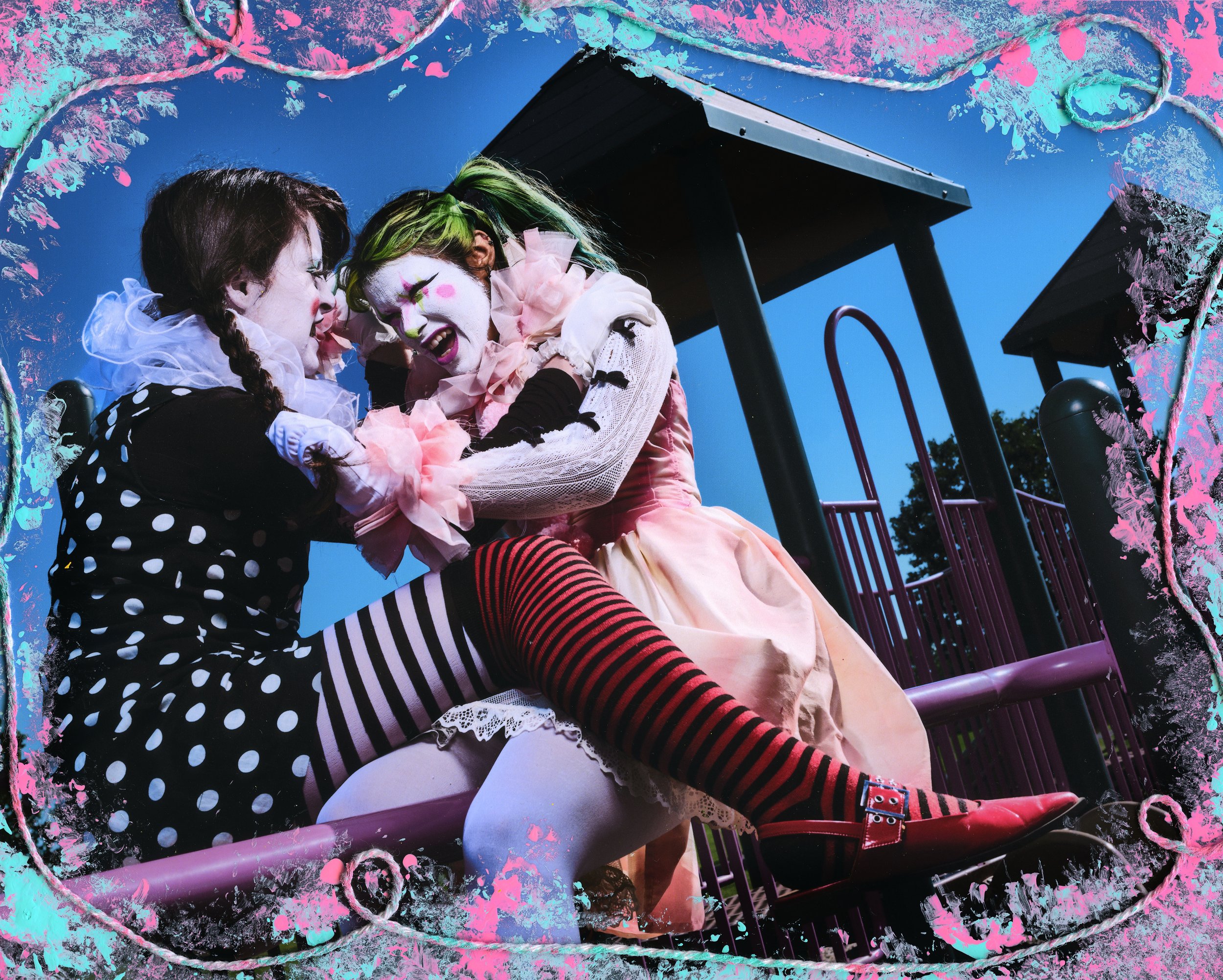 Two clowns dressed in colorful costumes sitting on a playground. One clown has black hair and the other has green hair, wearing striped stockings, and smiling while leaning against each other. The background shows a blue sky and playground equipment.