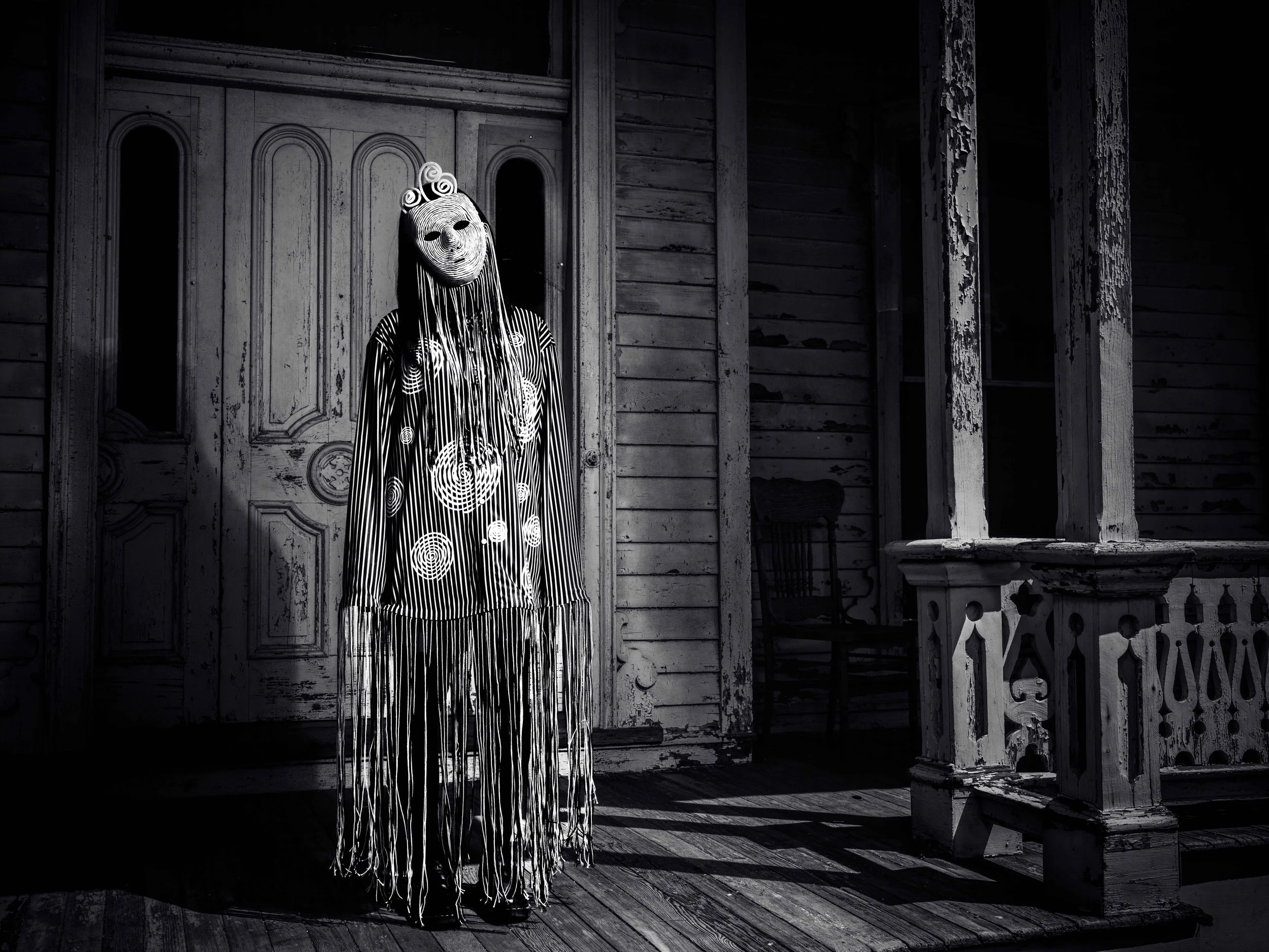 A person in a mask with a mask that looks like a human face, wearing a dress with a spiral circle pattern, standing in front of an old, weathered wooden house with a porch and door.
