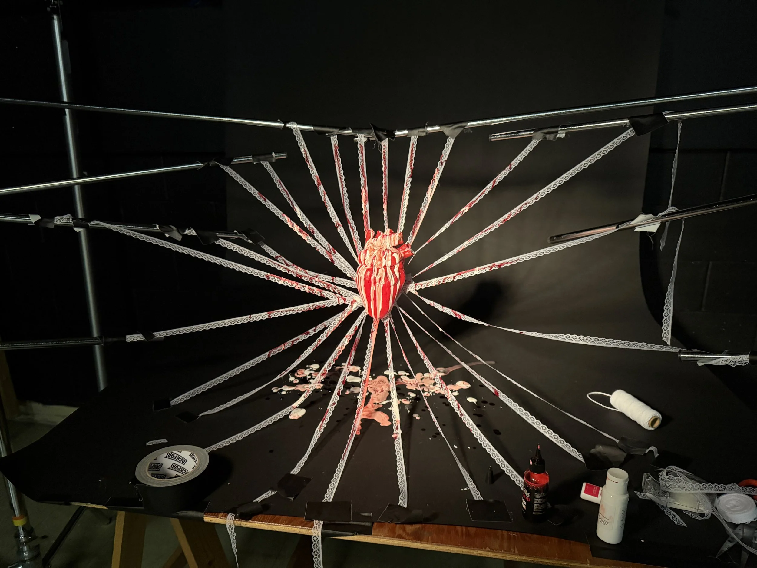 A large decorative spider with red and white striped body and white lace-like web marching across a black surface, with sewing supplies and tools scattered around.