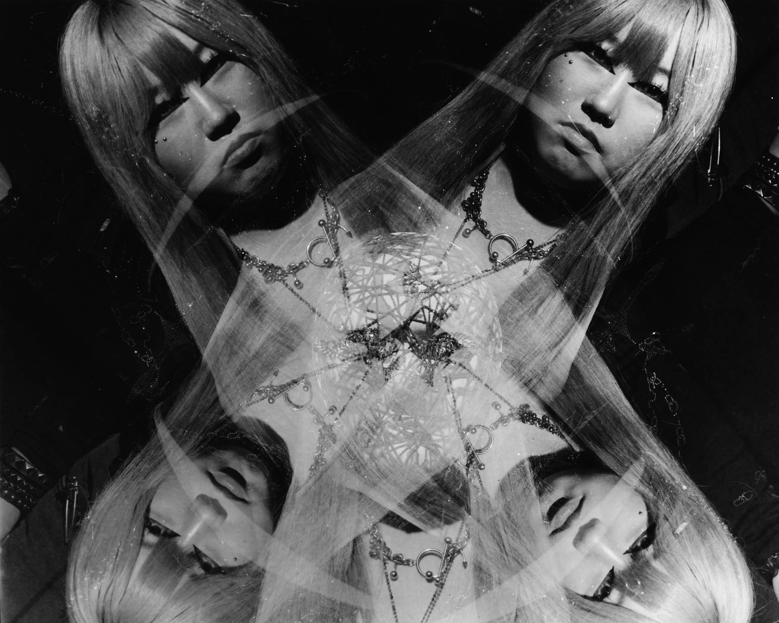 Black and white photo of three women with long hair wearing jewelry and clothing with metal spikes and studs, arranged in a triangular composition with reflections.